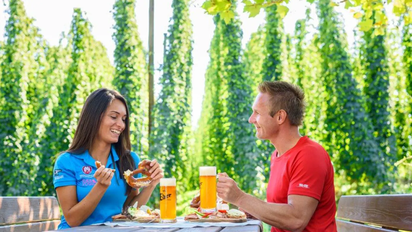 Zwei Gäste mit Bier und Brezen im Gastgarten vor hohen Hopfenranken